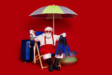 Photo of fat excited man pensioner dressed santa claus costume sitting deck chair enjoying vacation smiling isolated red color background © deagreez