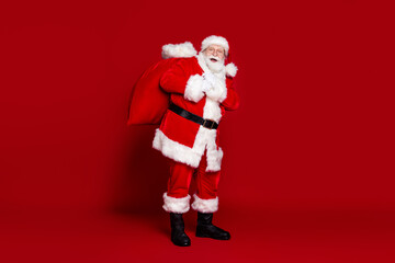 Photo of funny excited man pensioner dressed santa claus costume carrying big christmas sack smiling isolated red color background