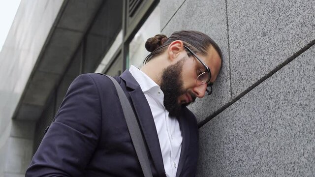 Deeply upset frustrated bearded man in eyeglasses leaning on wall of big office building, lost his job, male accountant lost lot of money and was fired from his job, redundancy or demotion, bullying