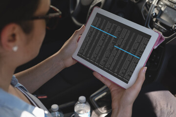 Biracial female programmer sitting in car, using tablet with coding on screen