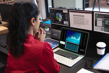 Biracial businesswoman sitting at desk using laptop with graph and statistical data on screen
