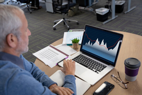 Caucasian senior businessman sitting at desk, using laptop with statistical data on screen