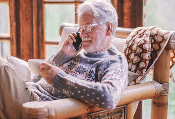 Smiling white haired senior man in winter sweater sitting in living room holding a tea cup using...