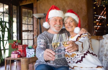 Fototapeta na wymiar Defocused senior couple in Santa hats hugging at home at Christmas time toasting with wine glasses. Sitting inside a rustic mountain chalet with Christmas tree, presents and lights on background
