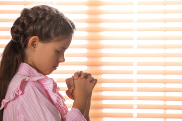 Cute little girl with hands clasped together praying near window. Space for text