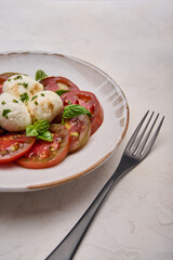 Vertical view caprese salad with red tomatoes, basil and mozzarella cheese in white plate and black fork