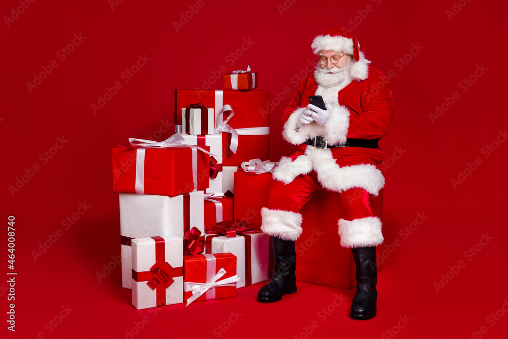 Wall mural Photo of pretty cheerful retired man wear santa claus costume spectacles preparing gifts typing modern device isolated red color background