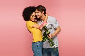 smiling man holding bouquet of roses while hugging happy hispanic woman isolated on pink.