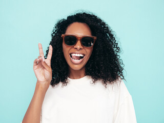 Beautiful black woman with afro  hairstyle. Smiling model in long t-shirt clothes. Sexy carefree female posing near blue wall in studio. Tanned and cheerful in sunglasses. Shows peace sign