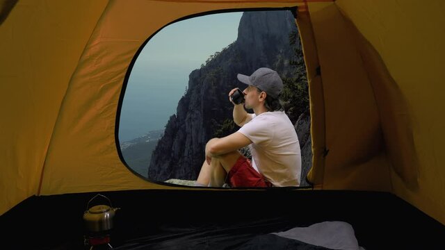 View from tent of hiker sitting and drinking tea on cliff and funicular going down from Ai-Petri mountain peak in background, Crimea