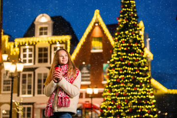 Christmas portrait of a beautiful girl in a warm scarf standing on the street, decorated for the New Year warming her hands with a deja paper cup with hot coffee