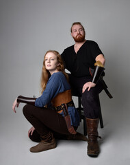 Fototapeta premium Full length portrait of red haired couple, man and woman wearing medieval viking inspired fantasy costumes, sitting poses, isolated on white studio background.