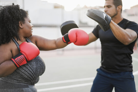 African curvy woman and personal trainer doing boxing workout session outdoor - Focus on left red glove