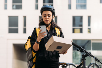Delivery woman in cycling helmet holding a cardboard box checking information on mobile phone - obrazy, fototapety, plakaty