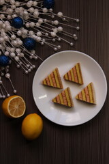 Christmas tree cookies on a white plate, decorated with white beads and blue Christmas balls and yellow lemons on a dark wooden background. New Year's still life with cookies, top view
