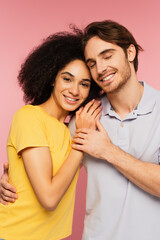 happy man with closed eyes hugging pretty hispanic woman looking at camera isolated on pink.