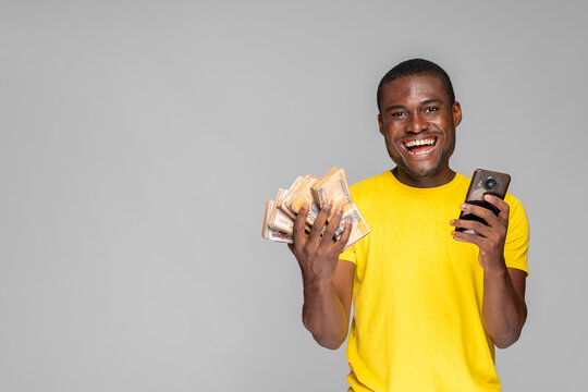 Excited Young African Man Holding Cash And Using His Phone Rejoicing