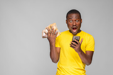 surprised looking young black man holding money and using his phone