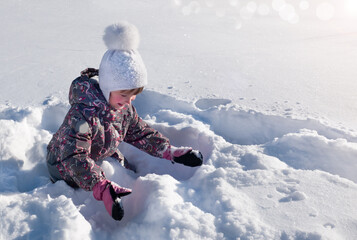 Fototapeta na wymiar A child sits in a snowdrift and plays with snow on a winter sunny day. Winter fun.