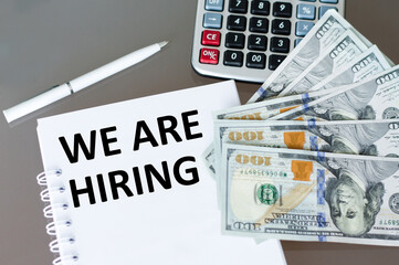 a white notebook with the text WE ARE HIRING on the table next to a pen, calculator and money. Business concept