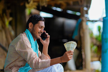 Young indian farmer showing money and talking on mobile phone at home