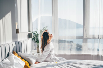  Woman in white bathrobe sitting near the big white window while stretching on bed after waking up.