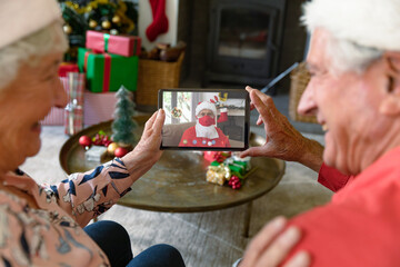 Caucasian senior couple on christmas video call on tablet with santa claus in face mask