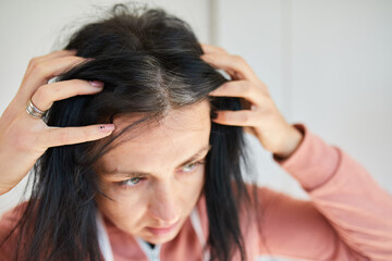 Fototapeta premium Portrait of a beautiful young woman examining her scalp and hair in mirror, hair roots, color, first grey hair, hair loss or dry scalp problem, or noticing that she is suffering from dandruff