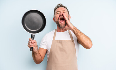 middle age handsome man feeling happy,giving a big shout out with hands next to mouth. frying pan concept