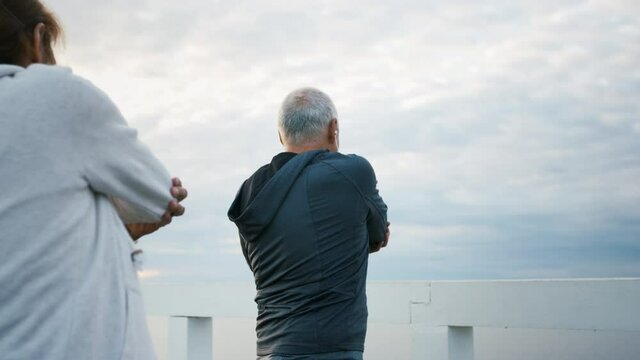 Senior couple outdoors on pier by sea at the morning, stretching after run.