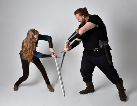 Full length  portrait of red haired  couple, man and woman wearing medieval viking inspired fantasy costumes, standing fighting pose holding  sword weapons, isolated on white  studio background.