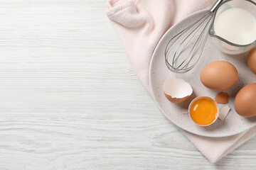 Chicken eggs, whisk and milk on white wooden table, top view. Space for text