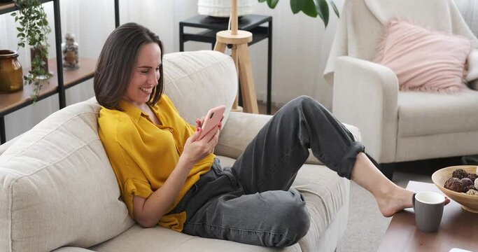 Happy woman having fun using mobile phone while relaxing on sofa at home