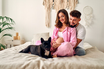 Young pregnant woman sitting on the bed in the bedroom with her husband, who hugs her tummy and with a family friend's dog. Maternity concept.