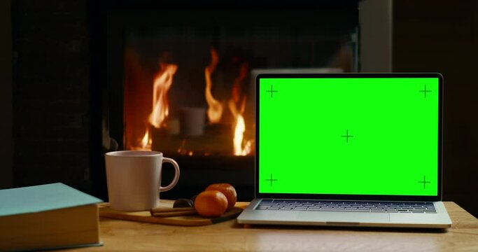 Laptop with Chromakey Green Screen on Table with Tea at Fireplace in Cozy Room or Winter Home Office. Christmas Online Shopping or Web site Background. 4K slider pan mockup shot