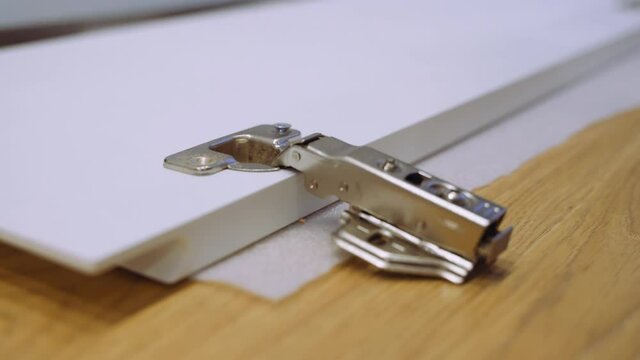 Carpenter man screws hinge to furniture door using electric screwdriver, hands closeup, side view. Carpenter is working making furniture in workshop.