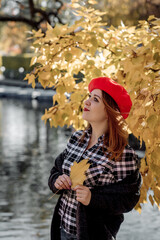 Beautiful caucasian adult young woman in a red beret hat stands near a pond in autumn during leaf fall and smiles emotionally