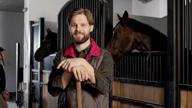 Bearded man holding shovel and smiling to the camera while working at the stable with horses at the background. Horse riding concept