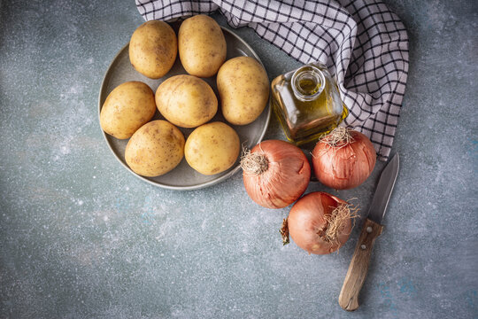 vista elevada de un plato con patatas, tres cebollas, una aceitera con aceite de oliva, un cuchillo y un trapo de cocina, sobre fondo azul claro y con espacio en blanco