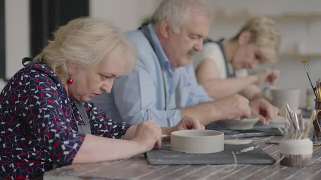 A group of elderly people at a master class in pottery together sculpt and cut a drawing on cups of clay for the manufacture of ceramic dishes