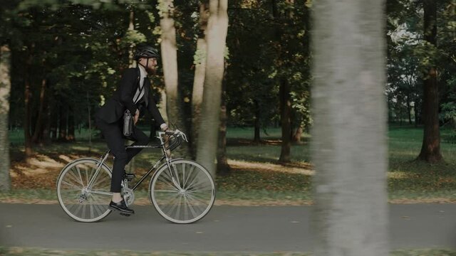 Businessman riding a bike in city park wearing helmet and a bag, side view. Businessman riding a bicycle in a public softwood park at the sunset. High quality 4k footage