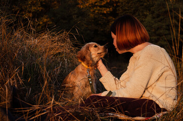 Woman with short red hair sitting on the ground in tall dry grass during sunset, looking at English cocker spaniel dog.