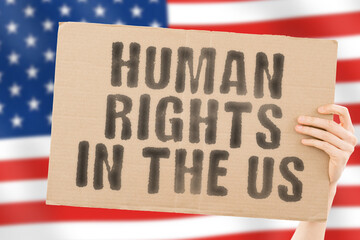 The phrase " Human rights in the US " on a banner in men's hand with blurred American flag on the background. Dictatorship. Equality. Freedom for people. Restrictions for civil citizens. Government