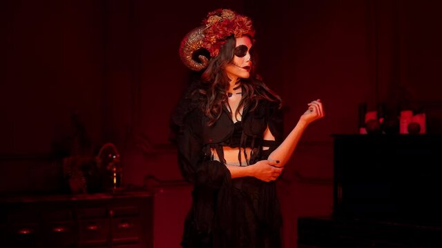makeup, halloween calavera catrina skeleton witch costume with horns, dried flowers, black torn clothes, black manicure on middle aged woman in red dark room, middle plan