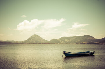 Pequeno barco ancorado sob as &aacute;guas calmas da enseada. Foto com tons esverdeados. Montanhas a o fundo.