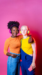 Two young diverse multicultural women best friends posing isolated background having fun laughing holding smartphone wearing wireless headphones