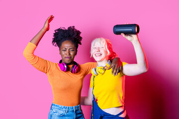 Young diverse black and caucasian women indoor holding wireless bluetooth speaker dancing together...