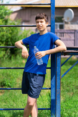 teenage boy exercising outdoors, sports ground in the yard, he stands near the horizontal bar, opens a bottle of water and drinks, healthy lifestyle