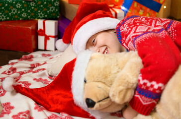 Fototapeta na wymiar Child girl sleeping in new year or christmas decoration. Holiday lights and gifts, Christmas tree decorated with toys. She's wearing a red sweater and a Santa helper hat