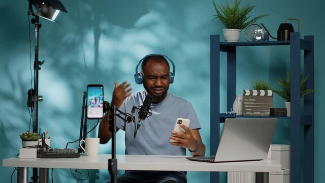 Frustrated vlogger giving bad news to subscribers on camera, using smartphone to film video for podcast. Blogger holding mobile phone while recording vlog on phone with gear in studio.
